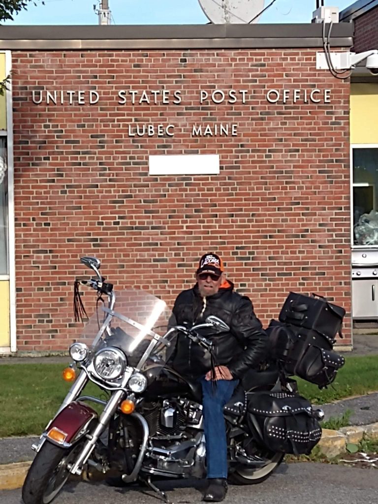 Man in a Laconia hat sitting on his bike in front of the Lubec, Maine post office