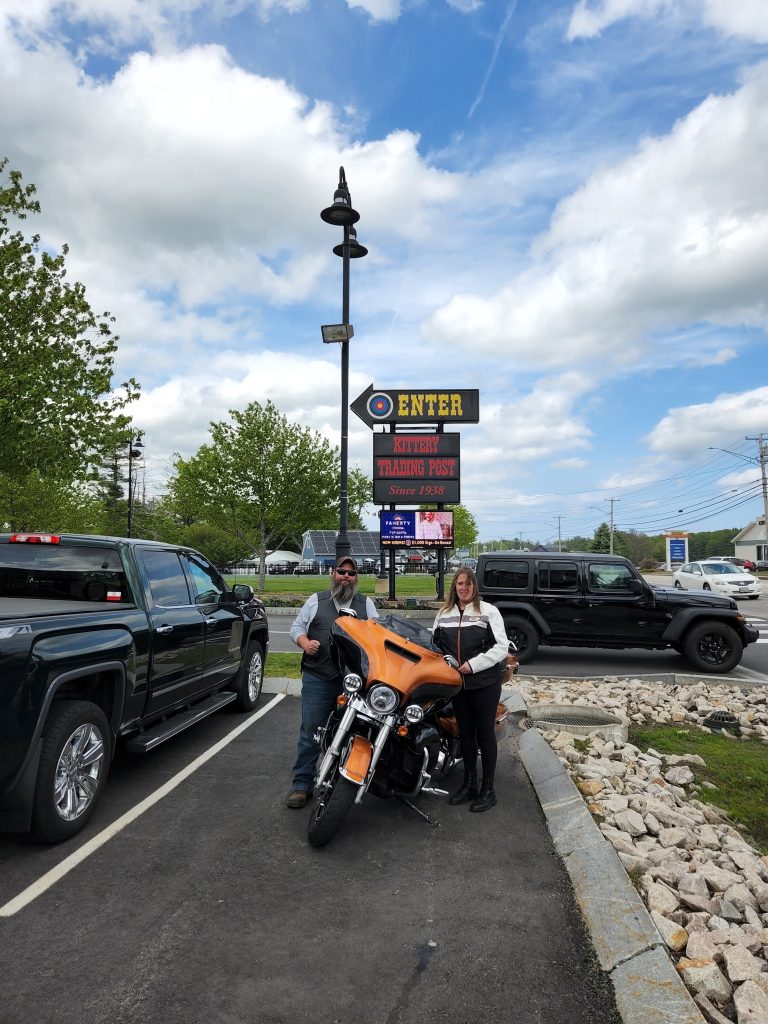Couple posing with their bike in front of the sign for the Kittery Trading Post
